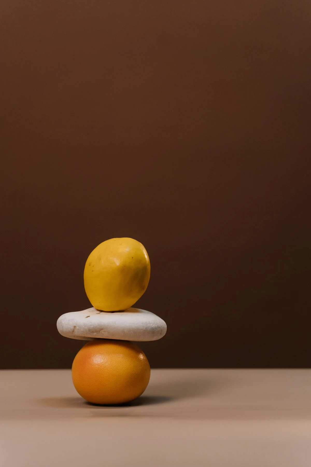 A lemon balanced above an orange with a pale oval stone between them against a warm brown background.