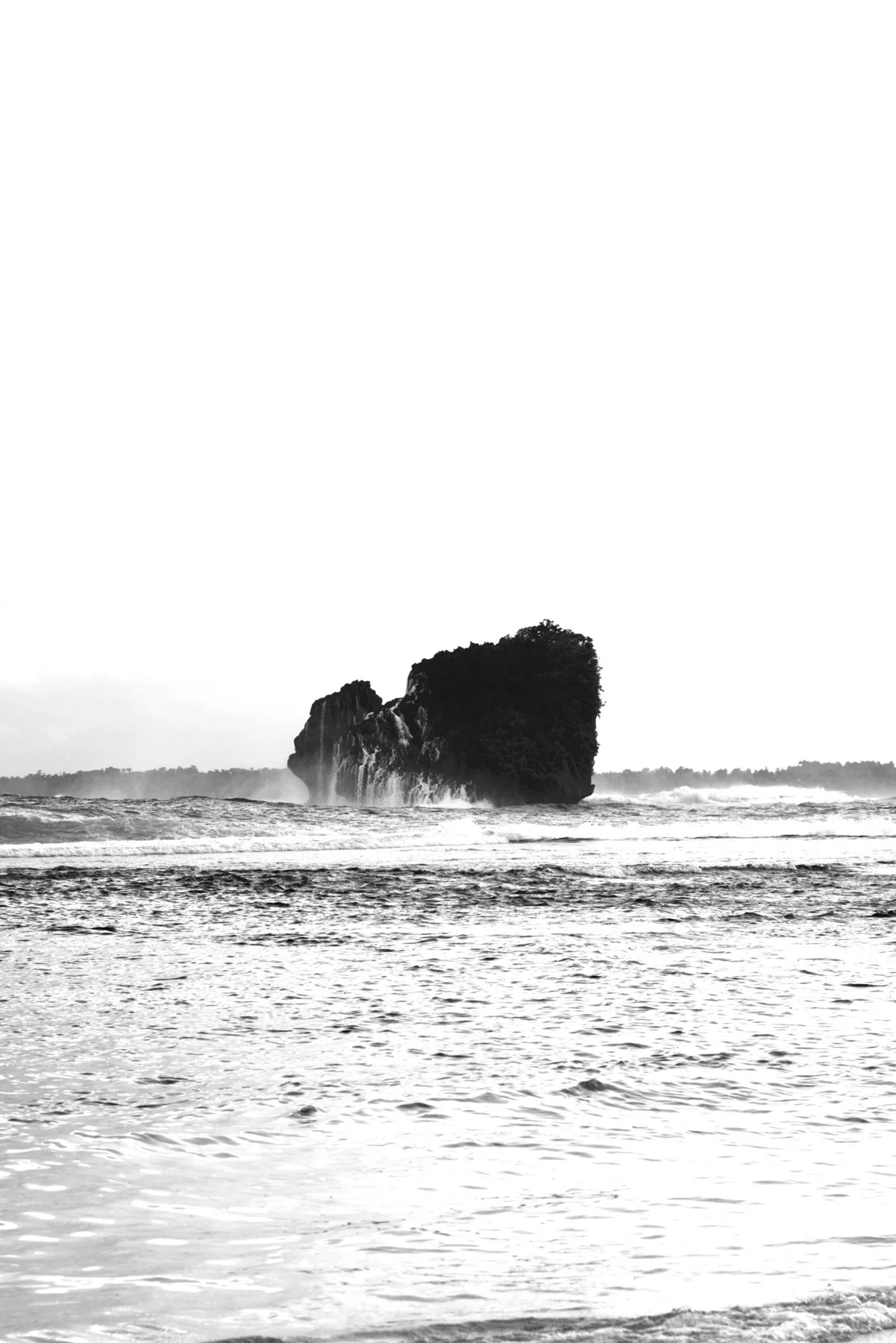 Black and white seascape with a dark offshore rock, breaking waves, and a broad pale sky.