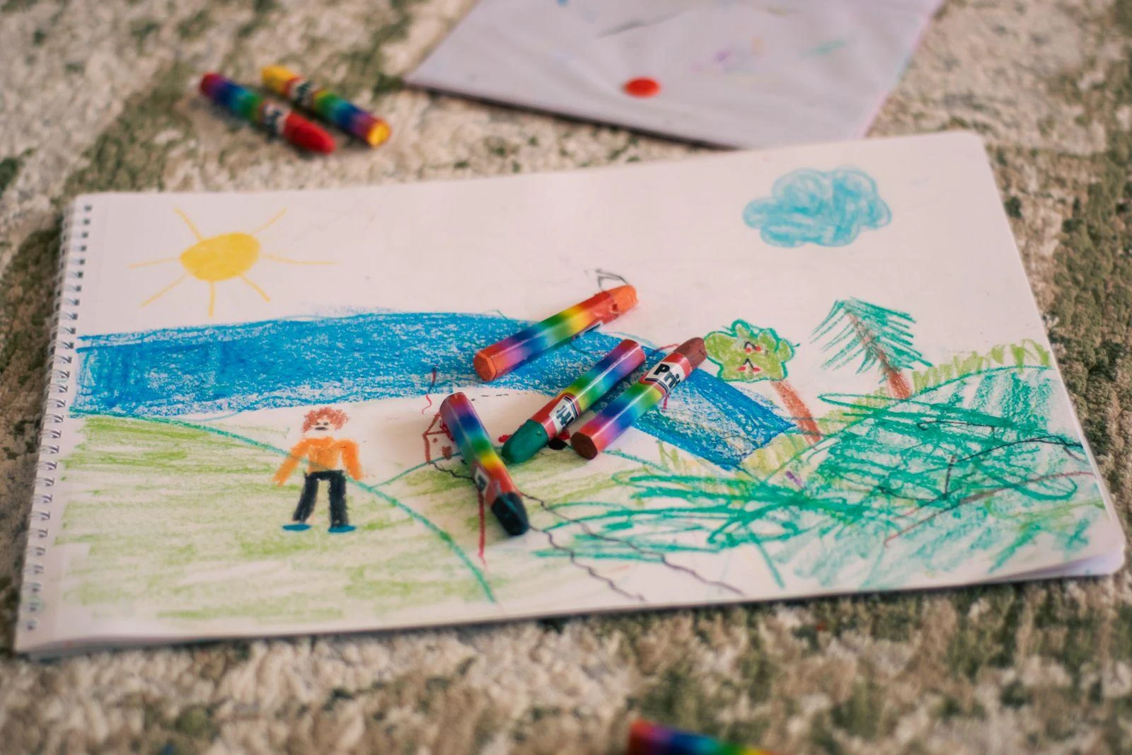 A colorful child’s landscape drawing lies on paper among crayons.